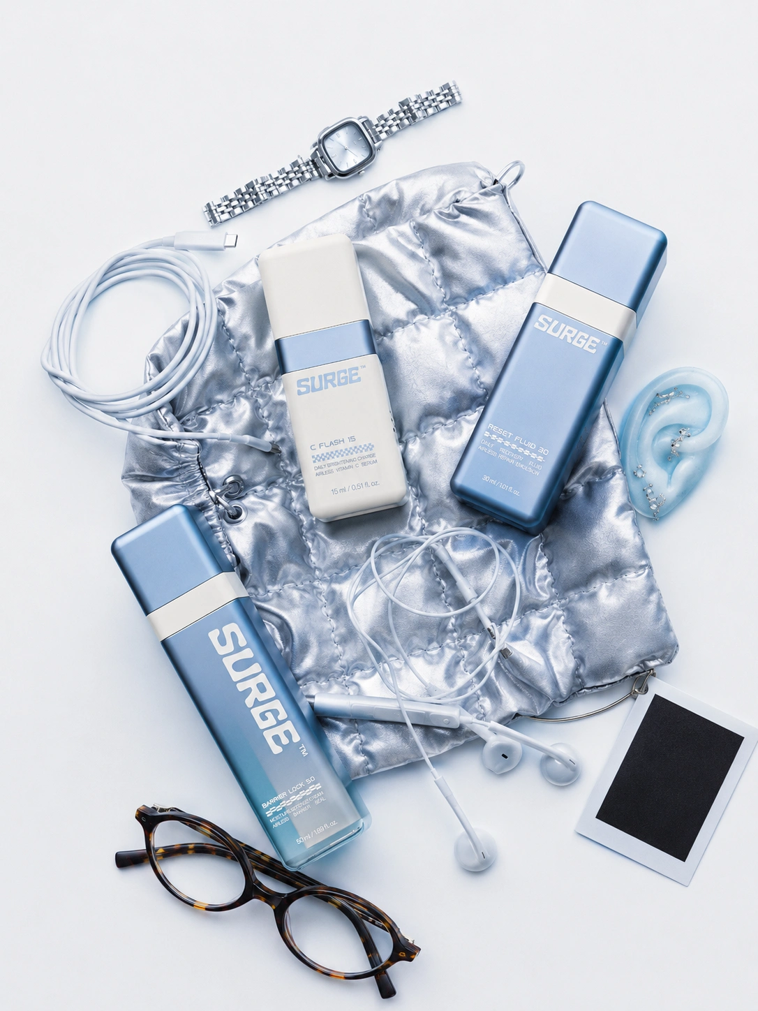 SURGE™ everyday-carry flat-lay — three bottles among a watch, earbuds and glasses on a silver pouch