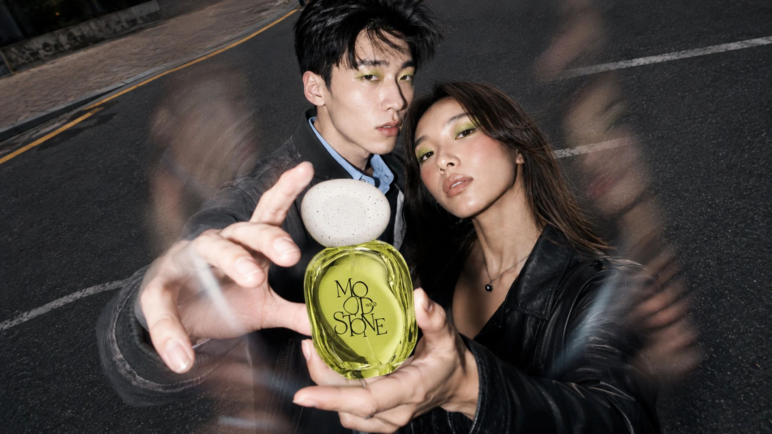 two models presenting a translucent crystal-green MOOD//STONE WILD perfume bottle in a nighttime street campaign image